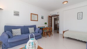 A living room with a blue sofa, a wooden coffee table, and a dining area with a table and chairs. - Cabanas T1 Apartment with Balcony & BBQ – 8min Walk to Beachfront (Faro)