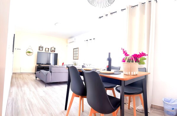 A modern living room with a grey sofa, a dining table with black chairs, a flat-screen TV, and a potted plant with pink flowers. - Villa Descansa with private Pool, Air-Conditioning, Sea View, Smart-TV & Wifi (Montaña Roja)