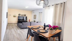 A modern living room with a flat-screen TV, grey sofas, a dining table with black placemats, and a potted plant. - Villa Descansa with private Pool, Air-Conditioning, Sea View, Smart-TV & Wifi (Montaña Roja)
