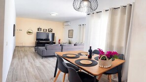 A modern living room with a flat-screen TV, grey sofas, a dining table with black placemats, and a potted plant. - Villa Descansa with private Pool, Air-Conditioning, Sea View, Smart-TV & Wifi (Montaña Roja)