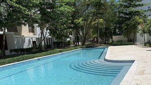 A rectangular swimming pool with a clear blue water surface, surrounded by well-maintained greenery and a tiled walkway. - Spacious apartment w/private rooftop and private cool water jacuzzi  (Punta Cana)