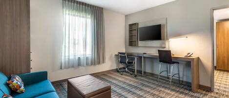 A hotel room with a blue sofa, a small ottoman, a desk with a computer and chair, a TV mounted on the wall, and a window with curtains.