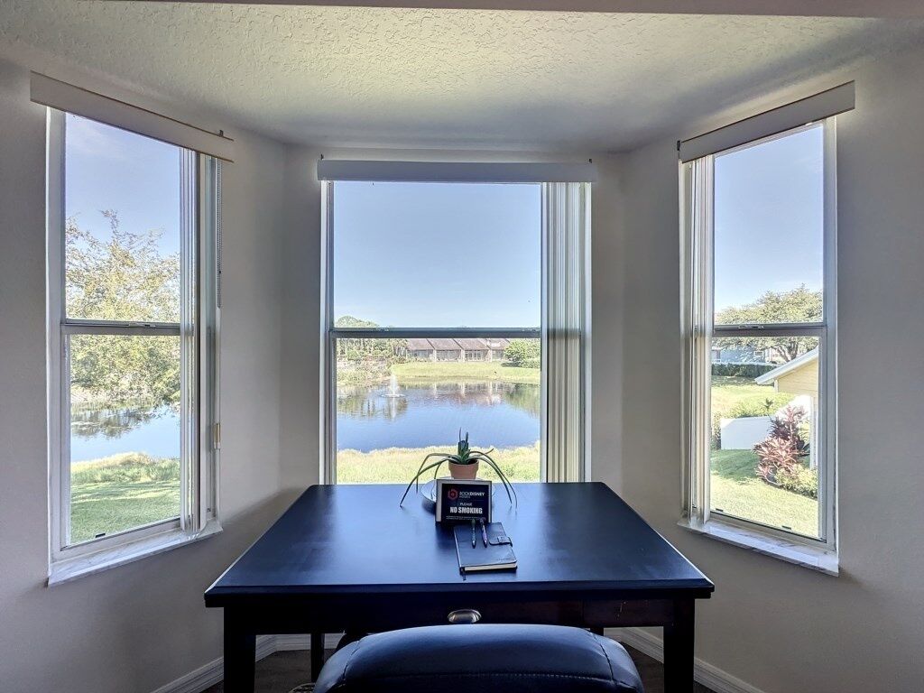 Quarto com escrivaninha e cadeira, janelas amplas com vista para um lago e vegetação, e um aviso de proibido fumar na escrivaninha.