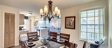 A dining area with a wooden table set for six, a chandelier, and a view of the kitchen and living area.