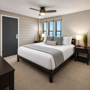 A bedroom with a bed, a ceiling fan, a window with a view, a nightstand with a lamp, and a door.