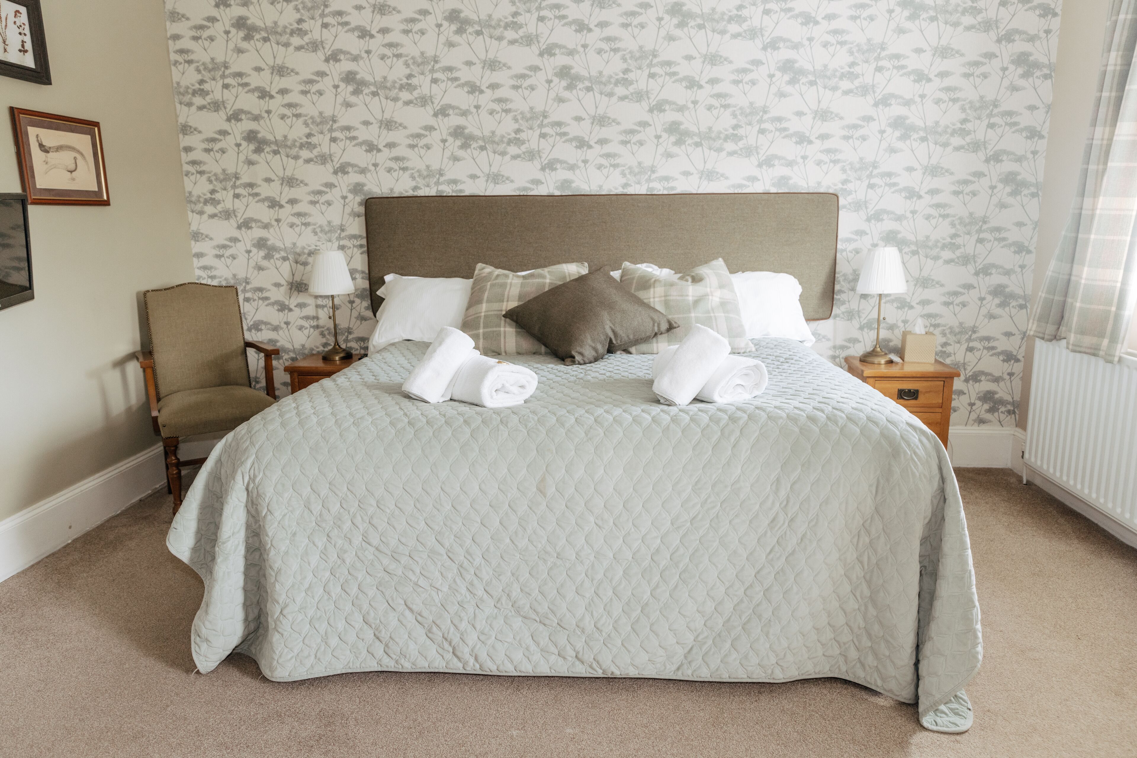 A neatly made bed with a quilted bedspread, a wooden nightstand with a lamp, and a chair.