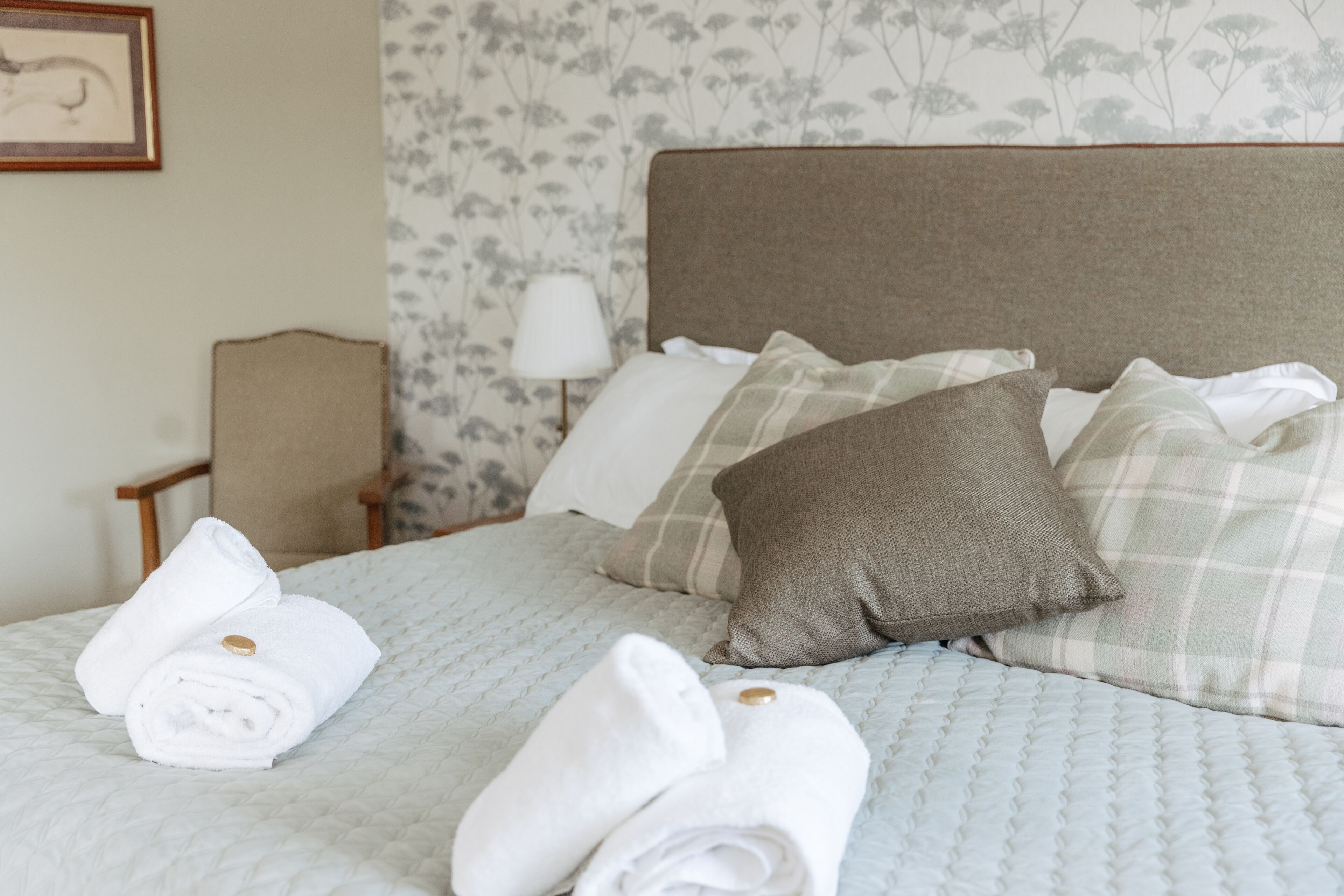 A neatly made bed with rolled towels and pillows, a lamp, and a chair in a room with floral wallpaper.