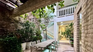 An indoor patio area with a long wooden table and chairs, surrounded by potted plants and a stone floor.