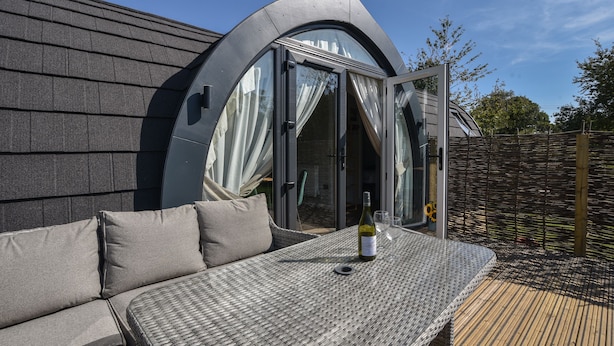 Een moderne buitenzitplek met een rieten tafeltje, kussens, een fles wijn en een glas.
