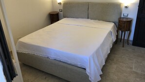 A neatly made bed with a white bedspread and lace trim, flanked by two bedside tables with lamps, in a room with beige walls and a tiled floor. - Bluehorizon (Torre Delle Stelle (Maracalagonis))