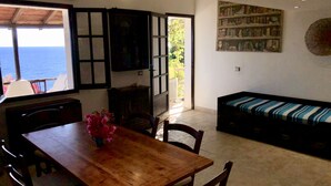A dining area with a wooden table and chairs, a sofa with a striped cushion, a bookshelf, and a view of the sea through a window. - Bluehorizon (Torre Delle Stelle (Maracalagonis))