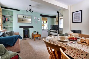 A cozy living room with a sofa, a dining table set for breakfast, a fireplace, and a bookshelf. - Otters Holt (Ambleside)