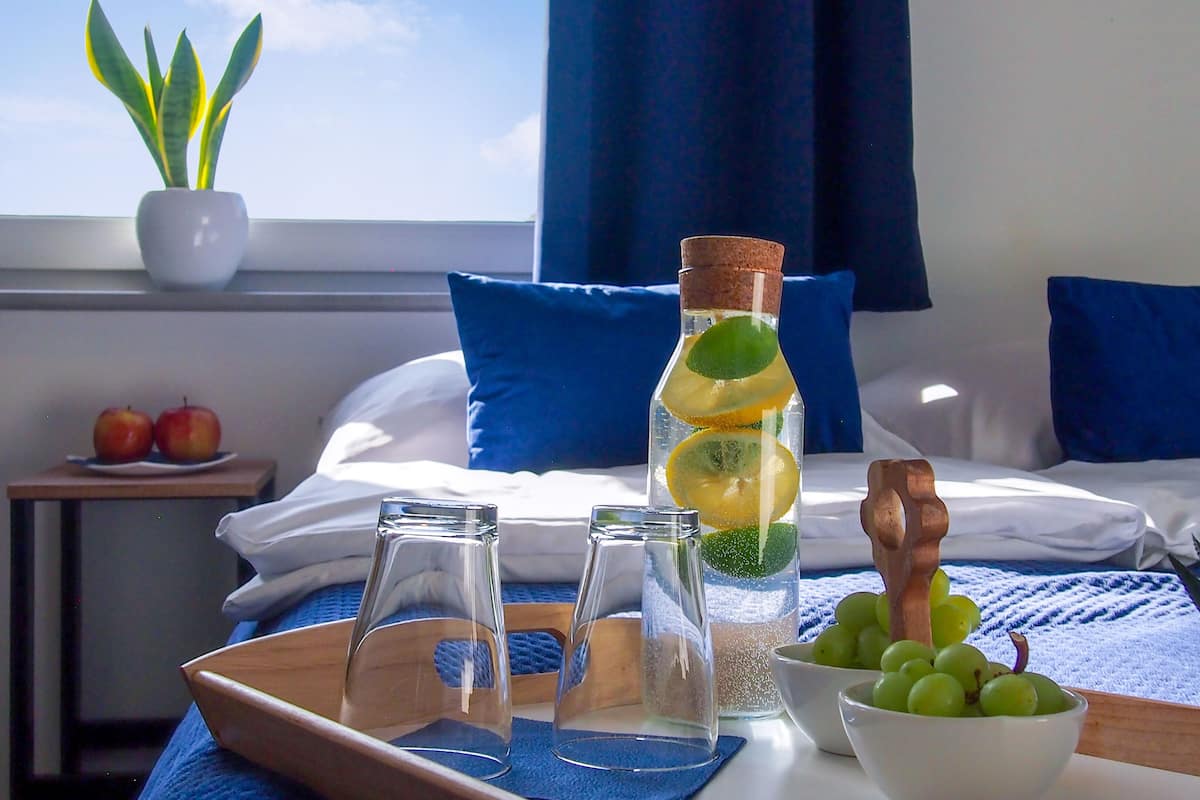 A hotel room with a bed, pillows, a window with curtains, a bedside table with apples, a tray with glasses, a bottle with lemon slices, a bowl of grapes, and a potted plant.
