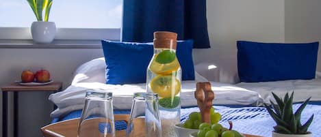 Une chambre d’hôtel avec un lit, des oreillers, une fenêtre avec des rideaux, une table de chevet avec des pommes, un plateau avec des verres, une bouteille contenant des tranches de citron, un bol de raisins et une plante en pot.