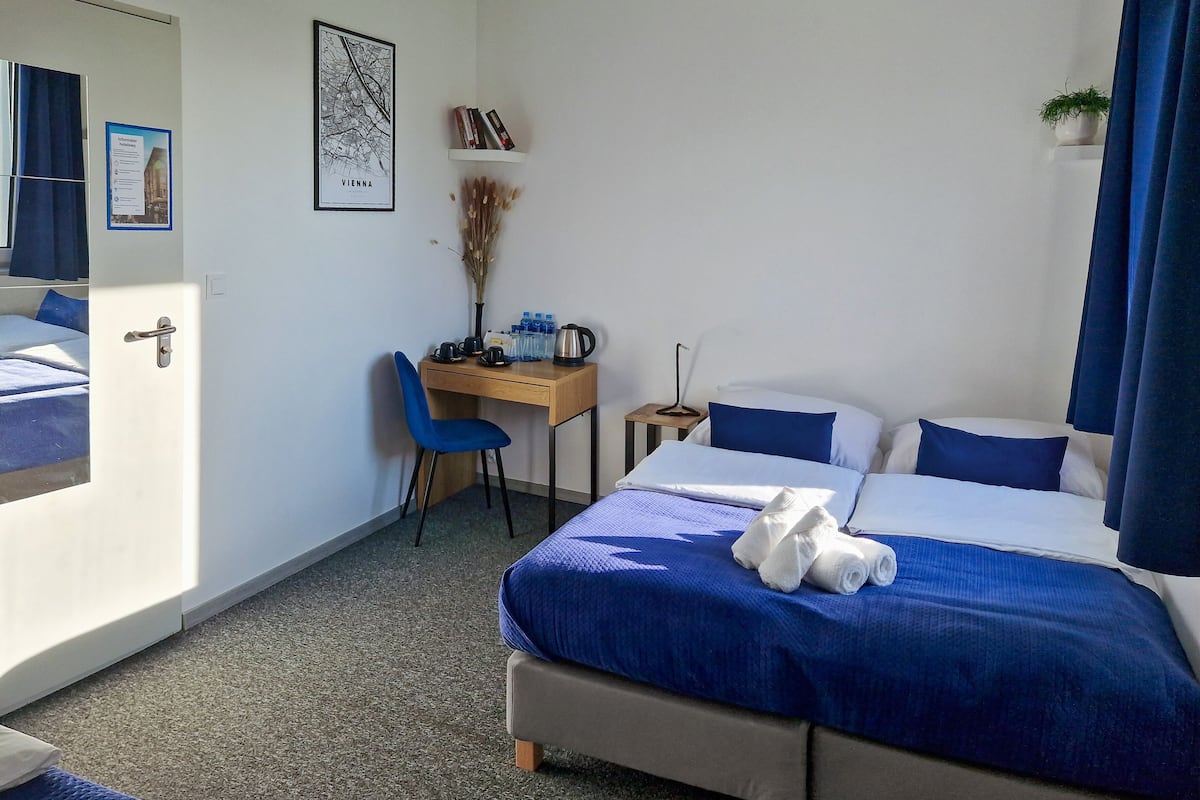 A hotel room with a bed, a desk, a chair, a small table, a potted plant, and a map on the wall.