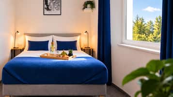 A neatly made bed with a blue blanket, a tray with fruits and a bottle, two bedside lamps, a framed picture on the wall, and a window with a view of trees.
