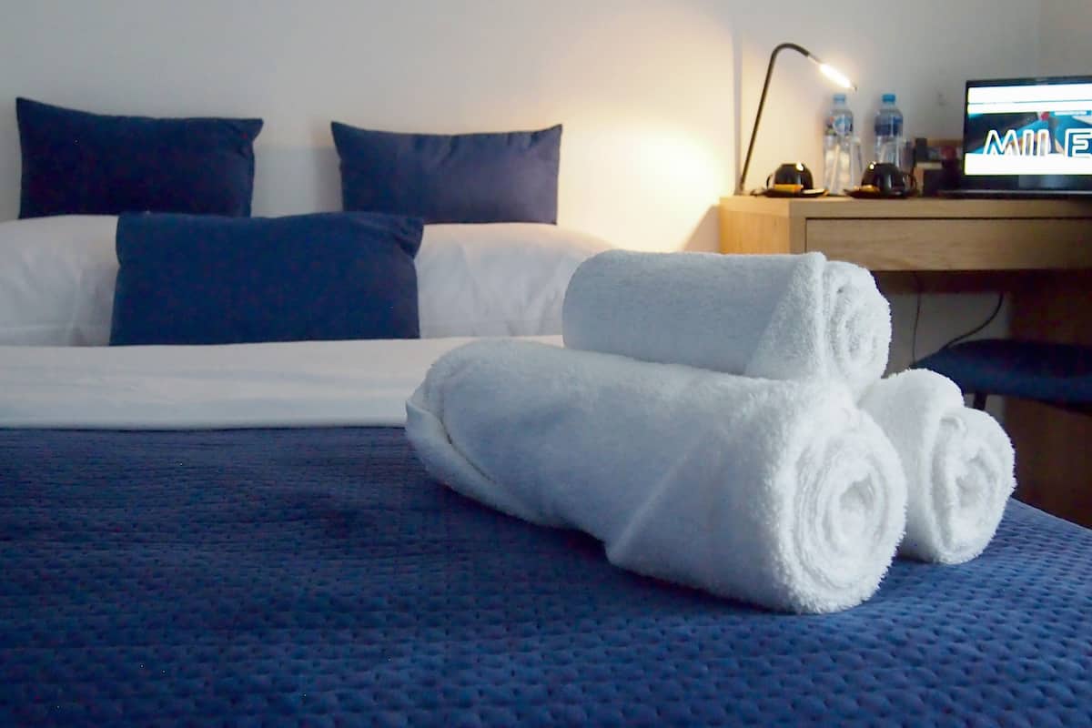 A neatly made bed with blue bedding, a pair of rolled towels, a desk with a computer and water bottles, and a chair.