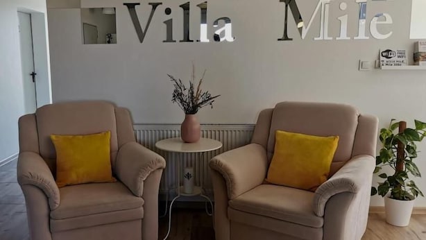 Une pièce avec deux fauteuils beiges, une horloge en bois et une enseigne murale portant l’inscription « Villa Mile ».