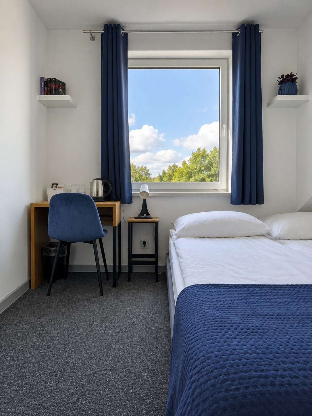 A hotel room with a bed, a desk with a chair, a small table with a lamp, a window with curtains, and a view of trees.