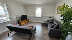 A modern living room with a grey sofa, a black sofa, a TV on a wooden stand, and a large window with a view. - Lakeview Court Southport (Southport)