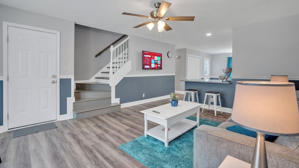 A modern living room with a staircase, a ceiling fan, a mounted TV, and a dining area with stools. - Spacious 4BR/2BA home ideal for families, contractors & extended stays. King primary + 3 queen BRs with Smart TVs & mini fridges. Fast WiFi, full kitchen, washer/dryer, backyard & deck. (Clarksville)