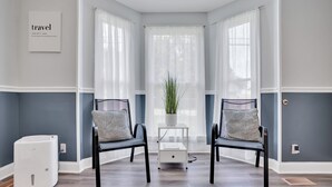 A room with two black chairs, a small white table with a plant, a white heater, and a framed 'travel' sign on the wall. - Spacious 4BR/2BA home ideal for families, contractors & extended stays. King primary + 3 queen BRs with Smart TVs & mini fridges. Fast WiFi, full kitchen, washer/dryer, backyard & deck. (Clarksville)