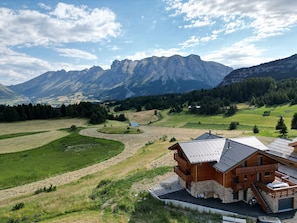 A wooden cabin with a balcony, surrounded by a lush green landscape and mountains in the background.