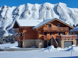 A wooden chalet with a stone foundation, wooden balconies, and a stone entrance, set against a snowy mountain backdrop.