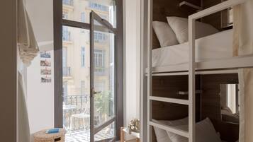 Una habitación con una cama litera, una ventana que da a edificios, una cesta de mimbre y una pequeña mesa.