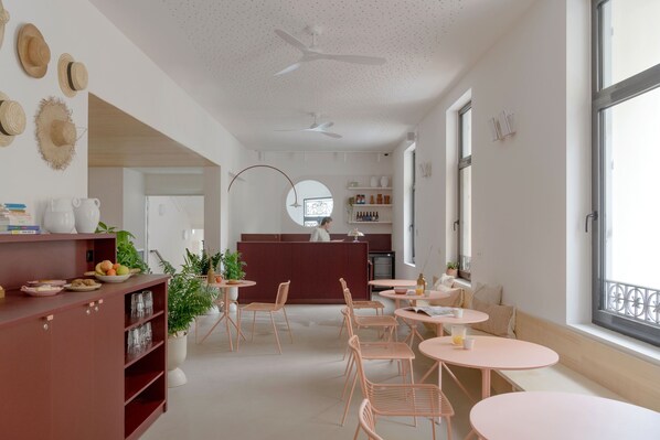 A modern dining area with round tables and chairs, a kitchen in the background, and large windows providing natural light. - Slo Hostel Nice (Nice)