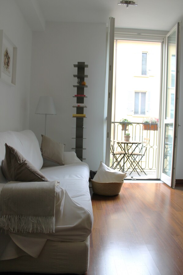 A modern living room with a sofa, pillows, a lamp, and a tall, narrow shelf. A balcony with a table and chairs is visible through an open door.