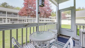 Un espace extérieur abrité, doté d’une table ronde en verre et de chaises blanches, d’une balustrade blanche et offrant une vue sur un bâtiment entouré d’une clôture blanche et agrémenté de feuillage rouge.