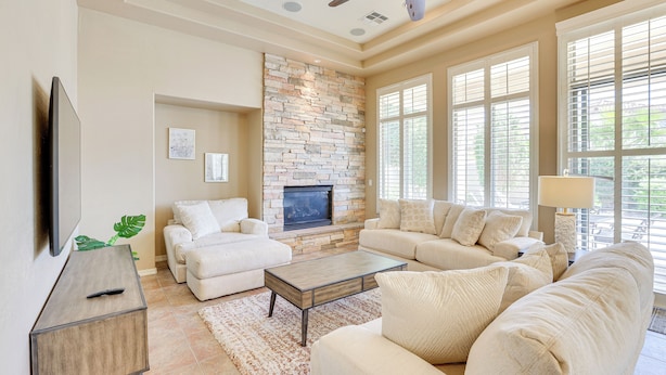 A living room with a stone fireplace, beige sofas, a coffee table, and large windows. - Private Pool & Patio at Home Near Goodyear (Litchfield Park)