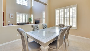 A dining area with a glass-top table, cushioned chairs, and a view of the living room through a large window. - Private Pool & Patio at Home Near Goodyear (Litchfield Park)