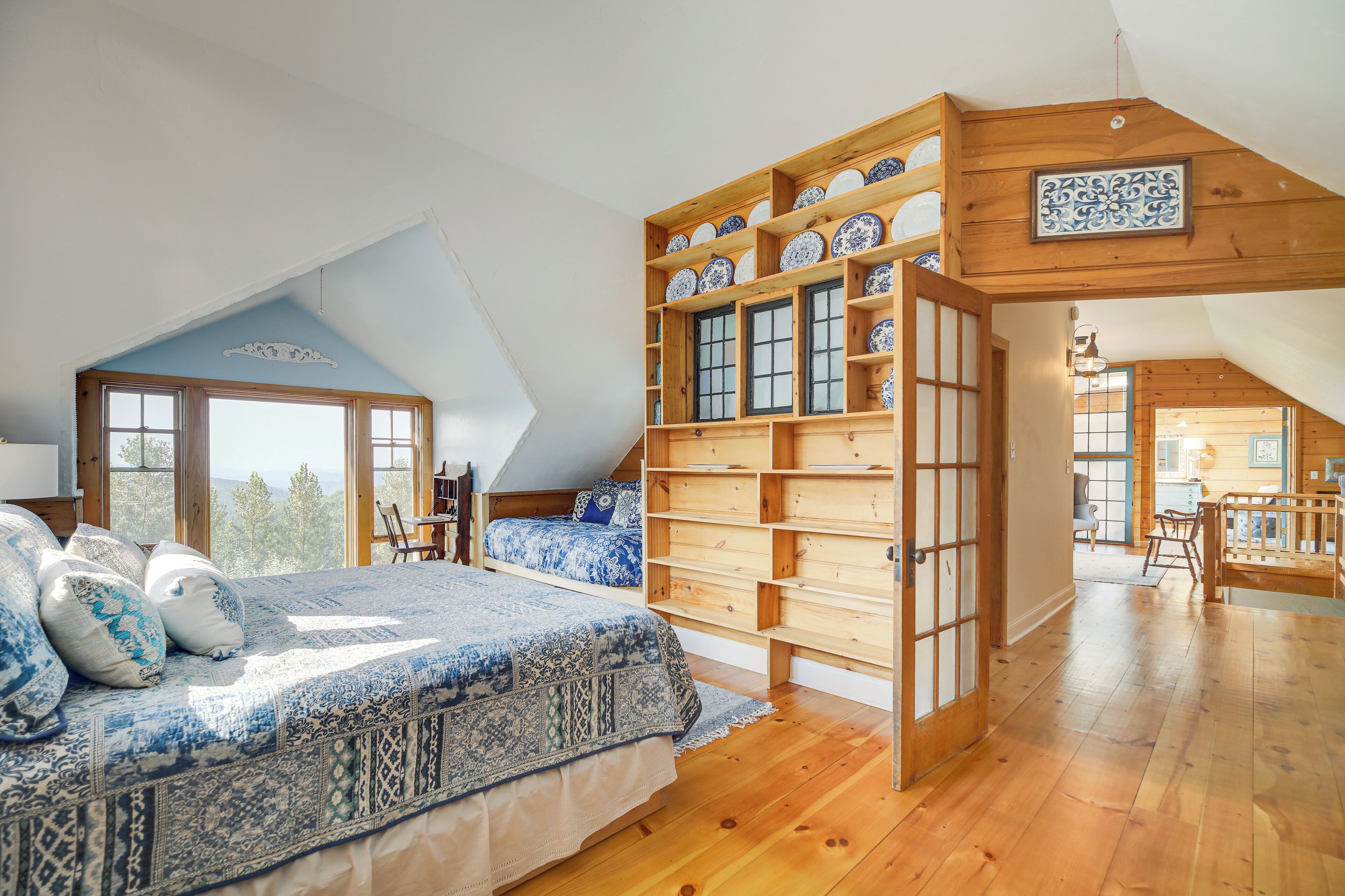 Ein Schlafzimmer mit schrägen Wänden, einem großen Bett mit gemusterten Textilien, Holzregalen mit dekorativen Tellern und Blick ins Freie durch ein Fenster.