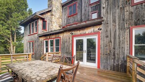 A wooden house with a stone chimney, a deck with a table and chairs, and a red-framed door. - Luxe 14-acre Vermont Countryside Lodge! (Washington)