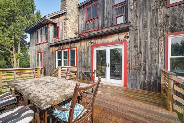 A wooden house with a stone chimney, a deck with a table and chairs, and a red-framed door.