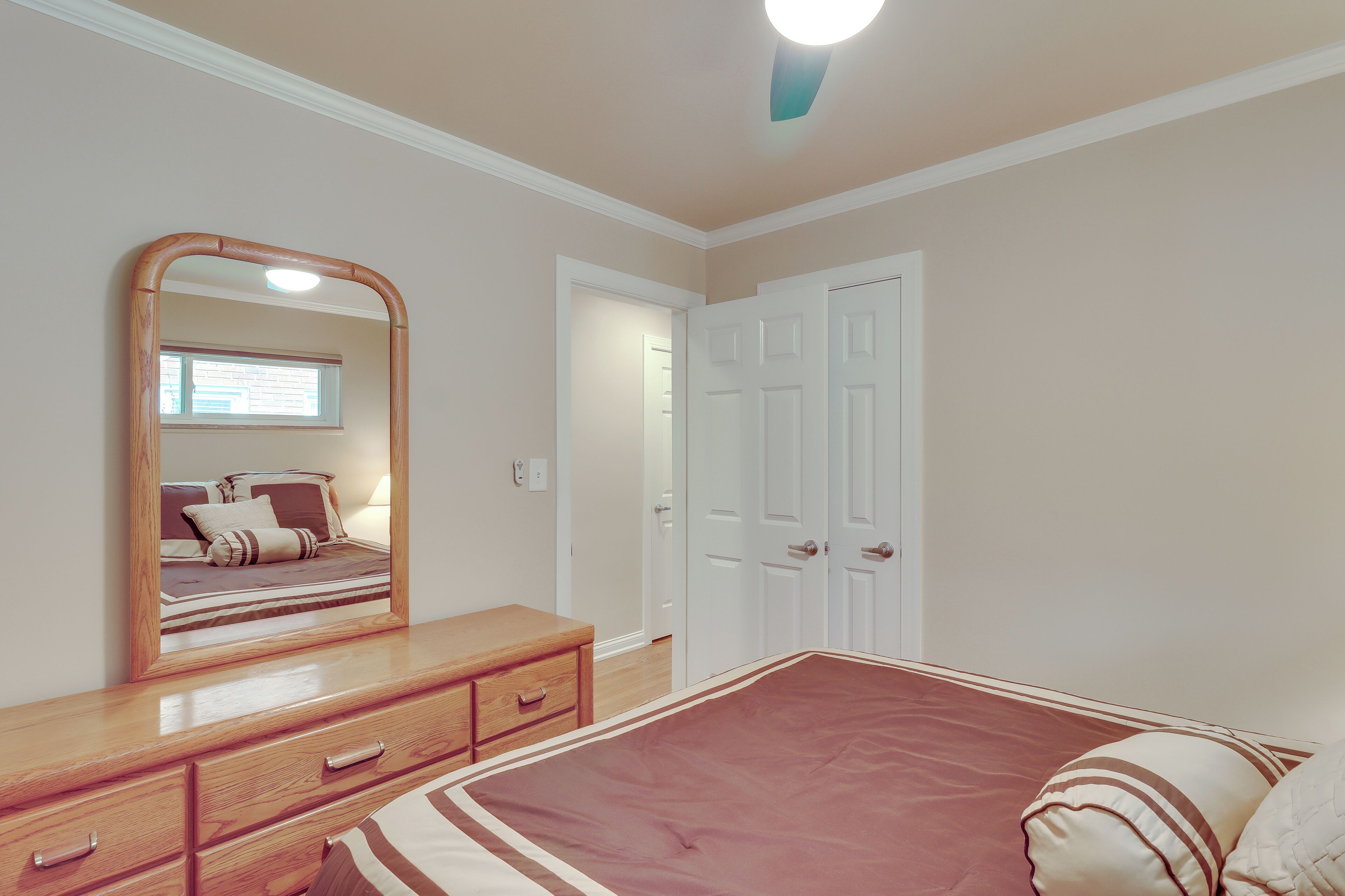 Ein Schlafzimmer mit einem Holzkapfchen, einem großen Spiegel, einem Bett mit roter Bettdecke, weißen Türen und einem Fenster mit Jalousien.