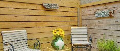 Una baita in legno con un cartello che dice 'Welcome to the Cabin', due sedie in metallo, un tavolo giallo in metallo con un vaso di girasoli e una pianta in vaso.