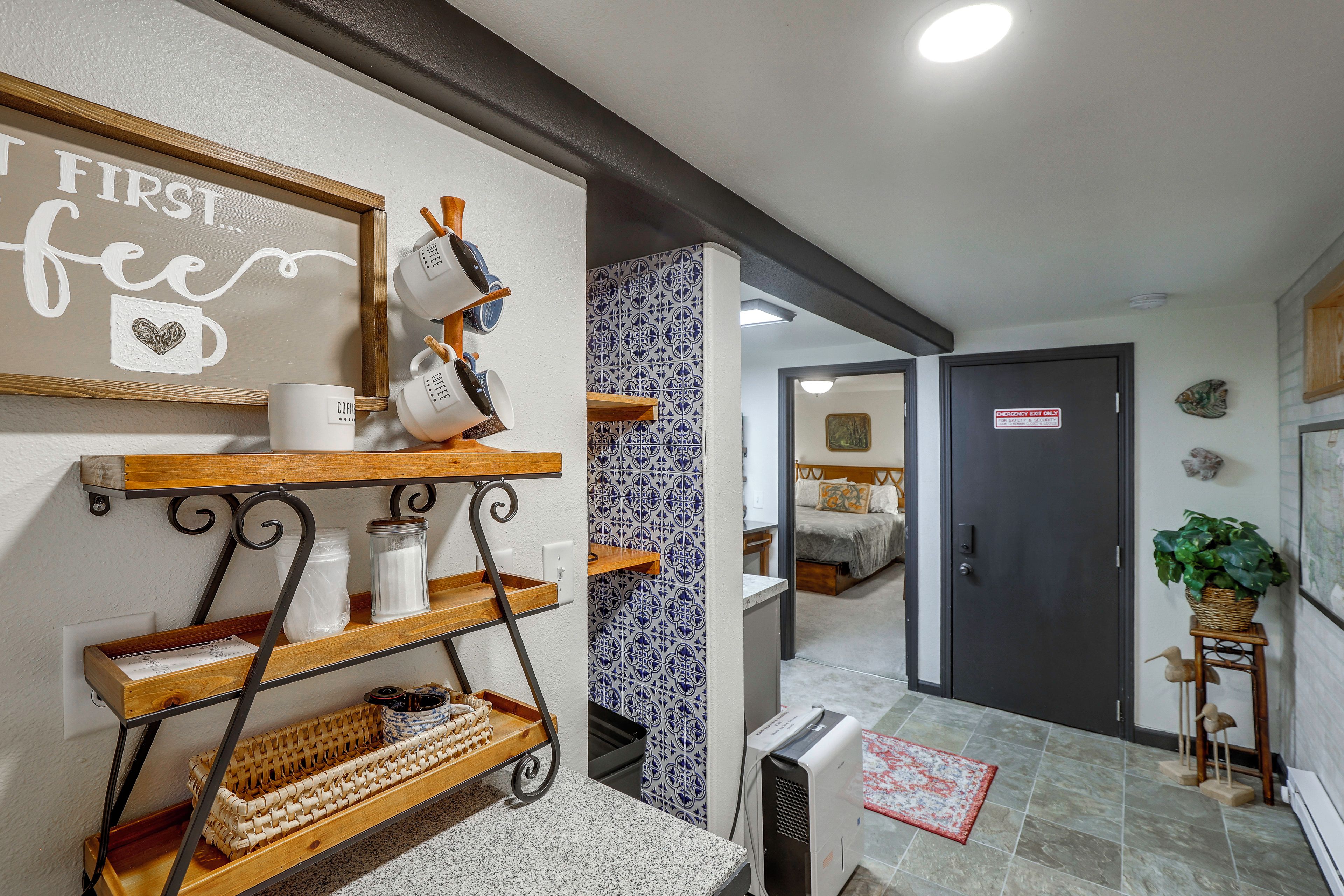 A hotel room with a bed, a small table, a shelf with mugs, and a wall with a sign.