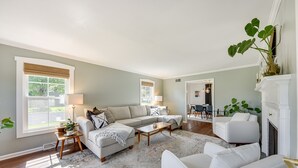 Uma sala de estar ampla, com um sofá grande, uma mesa de centro e uma lareira. Há duas janelas com persianas, um tapete e uma planta sobre uma mesinha lateral.