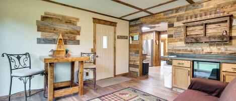 A rustic interior with exposed wooden beams, a wooden table with a chair, a checkered rug, and a kitchen area with wooden cabinets and a refrigerator.