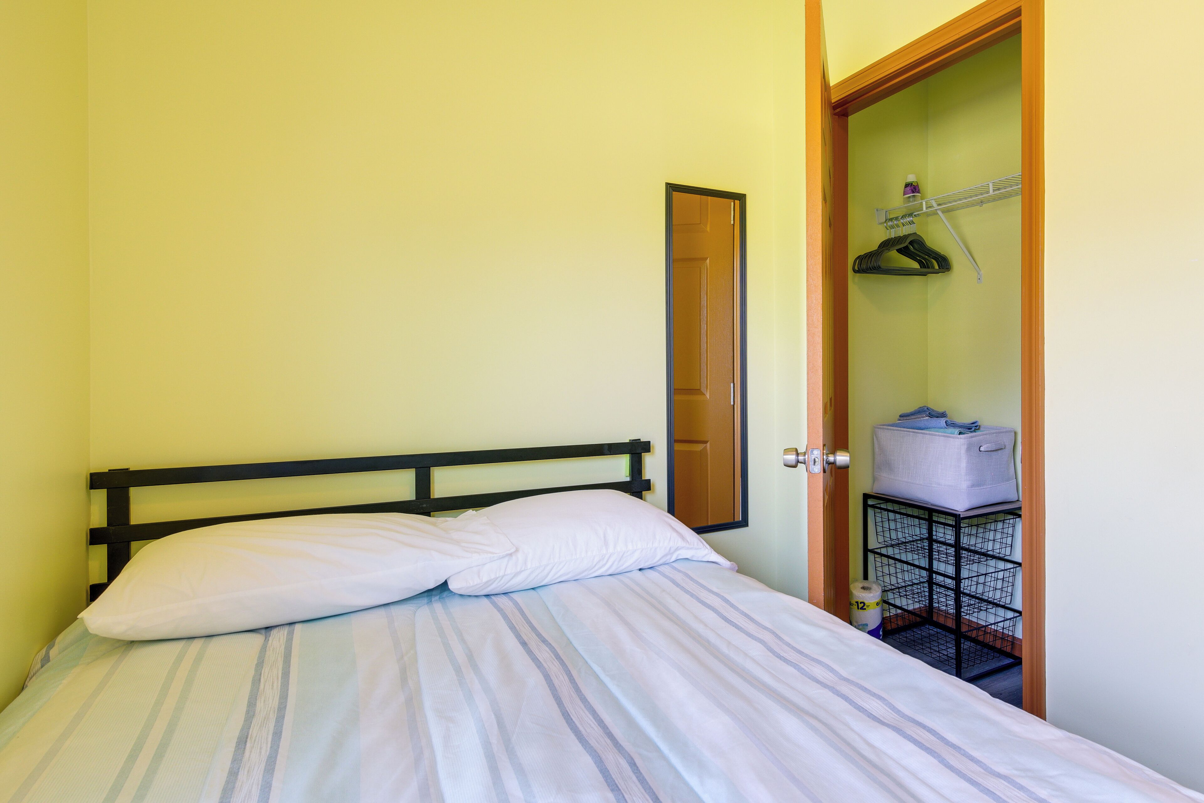 Un lit avec une tête de lit noire, un oreiller blanc et une couvre-lit à rayures. Une porte en bois se trouve sur la droite, donnant accès à un placard équipé d’une tringle et d’un bac de rangement.