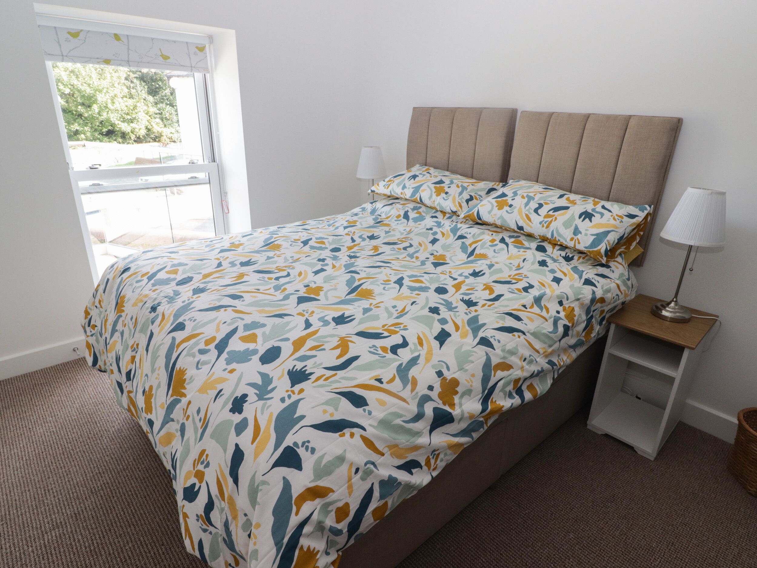 Quarto com cama, mesa de cabeceira, luminária e janela com vista para vegetação.