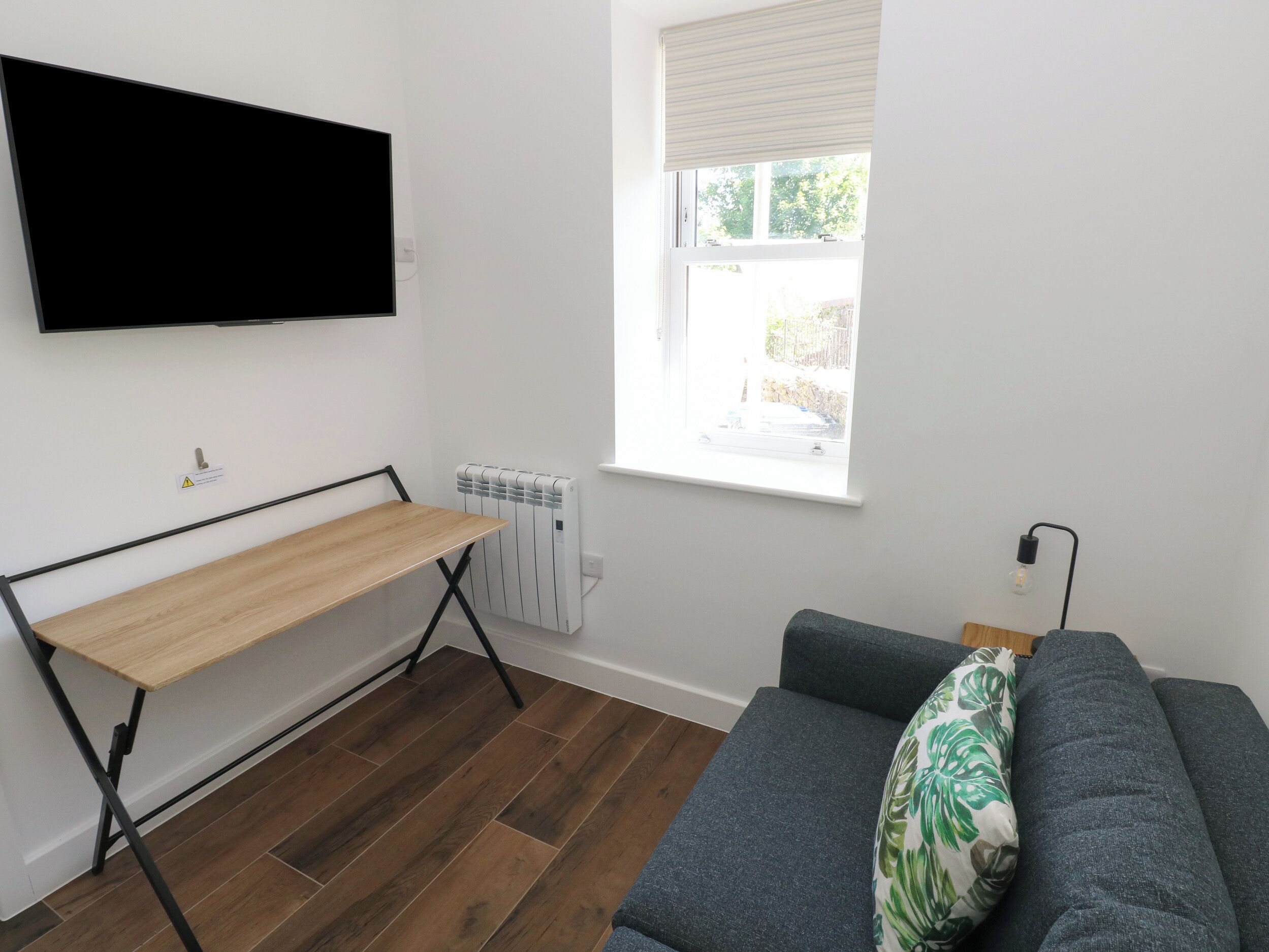Ett rum med ett skrivbord i trä, en grå soffa, ett element och ett fönster med persienner.