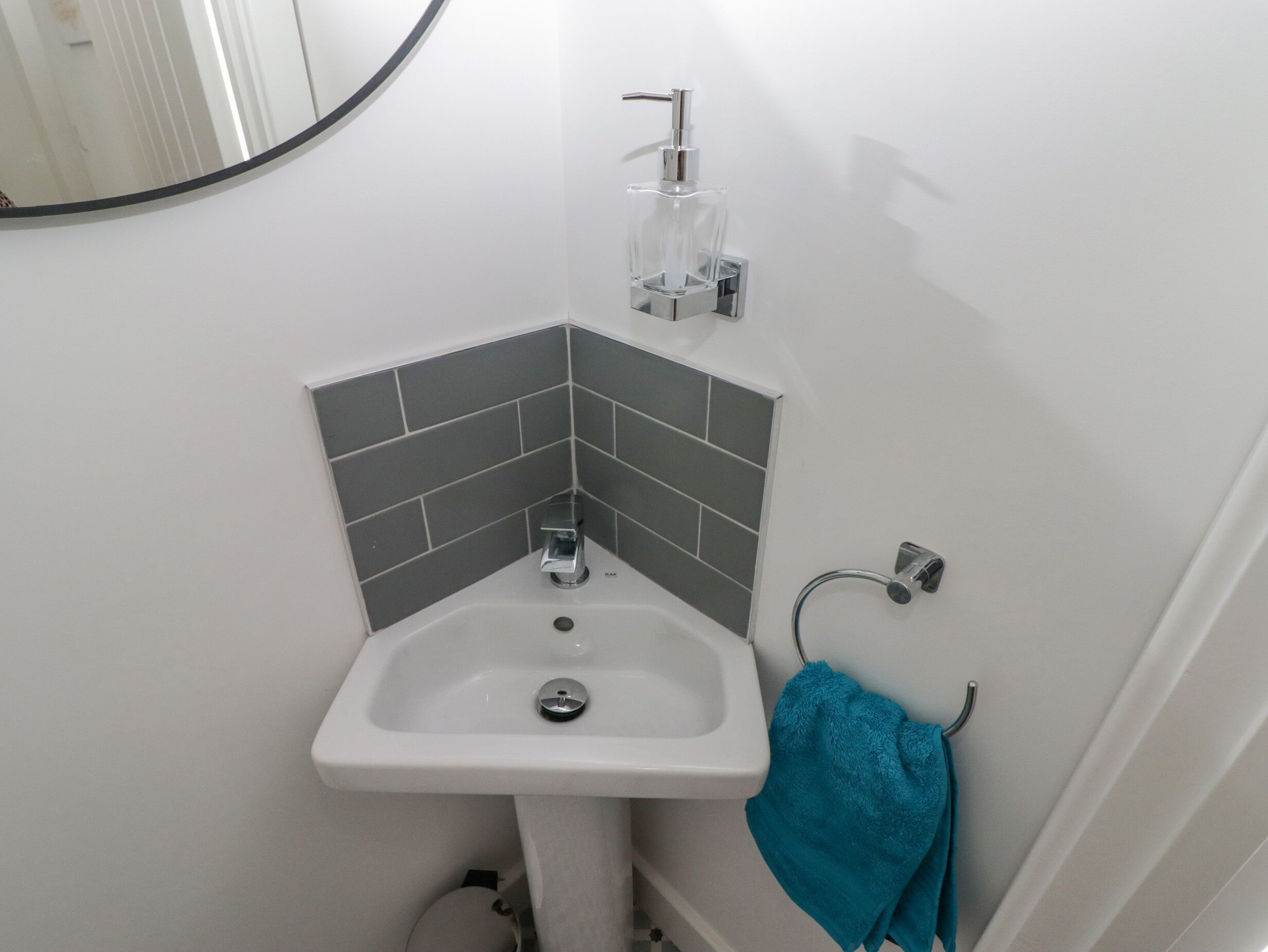 Ein Badezimmer im Eckbereich mit einem weißen Waschbecken, grauen Fliesen, einem Glas-Seifenspender und einem türkisfarbenen Handtuch an einem Metallhaken.
