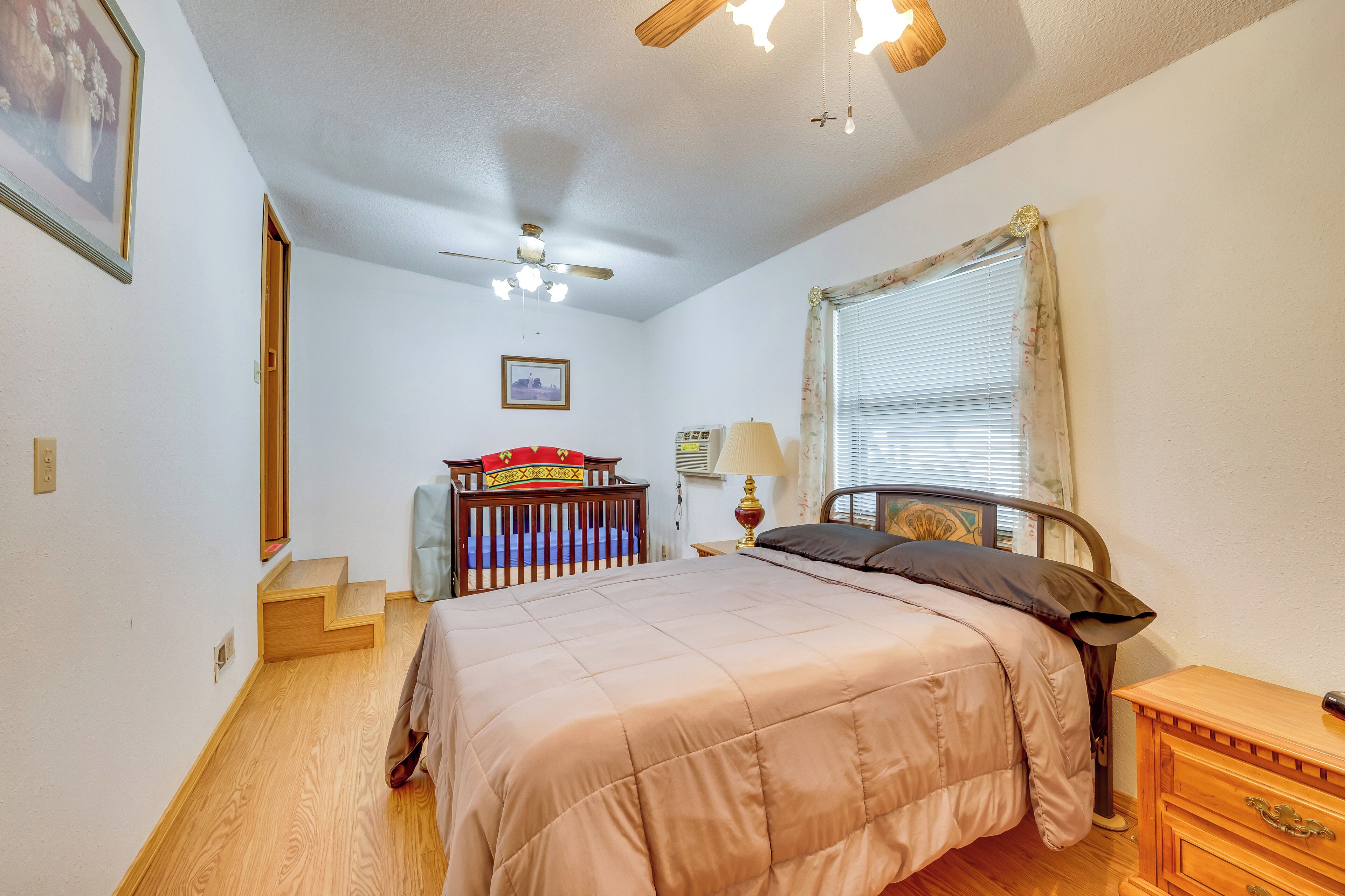 A bedroom with a bed, wooden dresser, nightstand, ceiling fan, and a window with blinds.