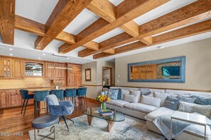 A spacious living area with a large sofa, a coffee table, and blue chairs. There is a kitchen with wooden cabinets and a dining area with bar stools.