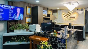 Una reception hotel con uno schermo digitale che mostra una lista di canzoni popolari, un'area di accoglienza con un bancone in marmo e un cartello dell'hotel.