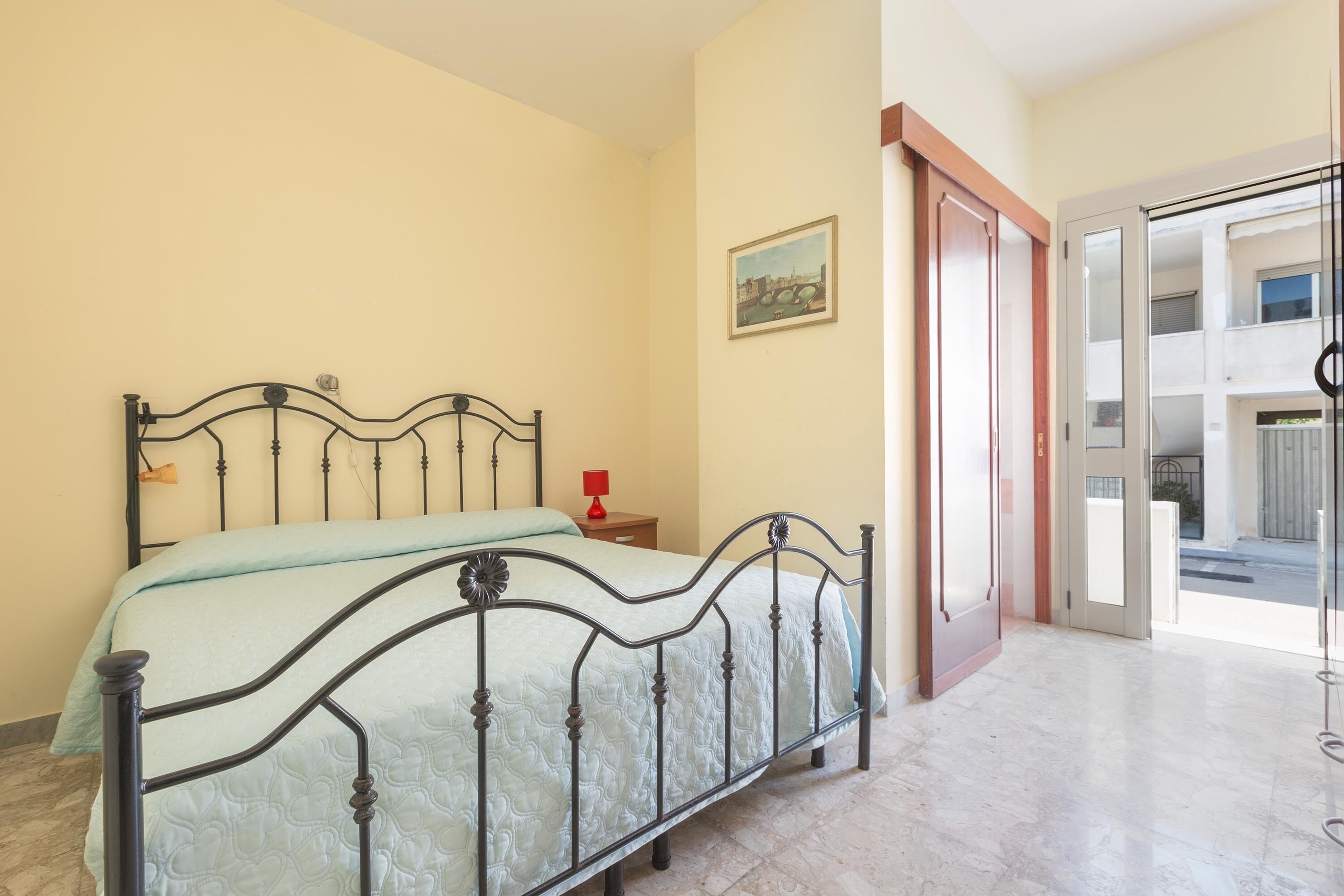 A bedroom with a metal bed frame, a bedside table with a red lamp, and a door leading to a balcony.